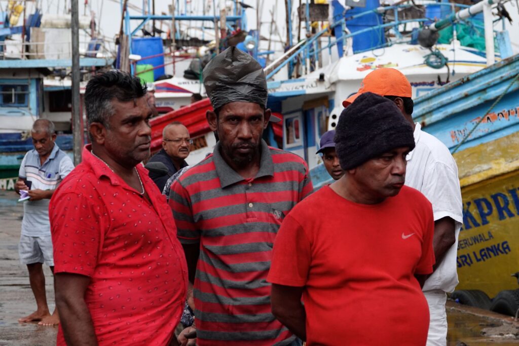 Drei Fischer in roter und gestreifter Kleidung stehen am Hafen von Beruwala, im Hintergrund traditionelle Fischerboote mit langen Angelruten und das Schild "Beruwala Sri Lanka"