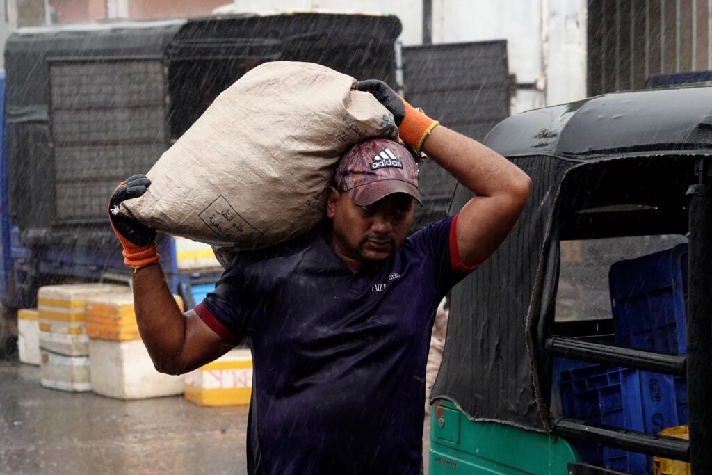 Marktarbeiter trägt schweren Sack im strömenden Regen auf dem Fischmarkt Beruwala, Sri Lanka, mit sichtbaren Regentropfen und Tuk-Tuk im Hintergrund