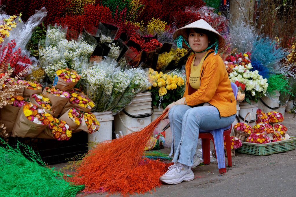 Junge Blumenhändlerin mit traditionellem Vietnamesenhut zwischen bunten Blumen auf dem Đà Lạt Markt