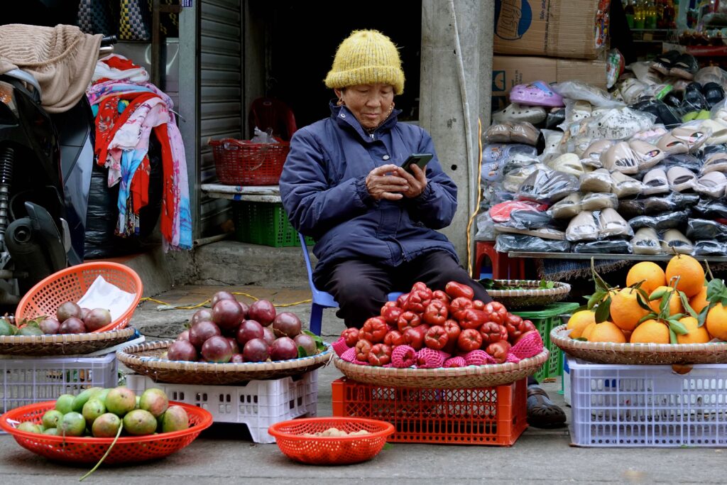 Obsthändlerin mit Smartphone auf dem Đà Lạt Markt umgeben von bunten Früchten in Körben